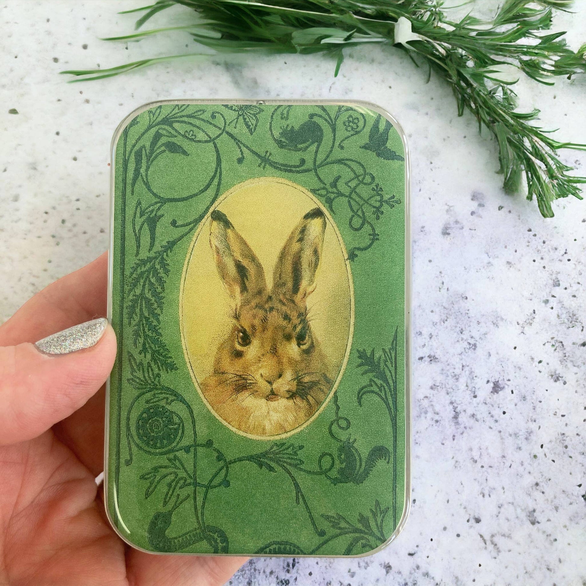 Green tin with rabbit illustration held by a hand on a textured surface with greenery.