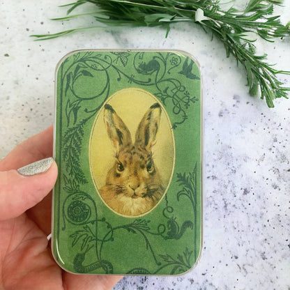 Green tin with rabbit illustration held by a hand on a textured surface with greenery.