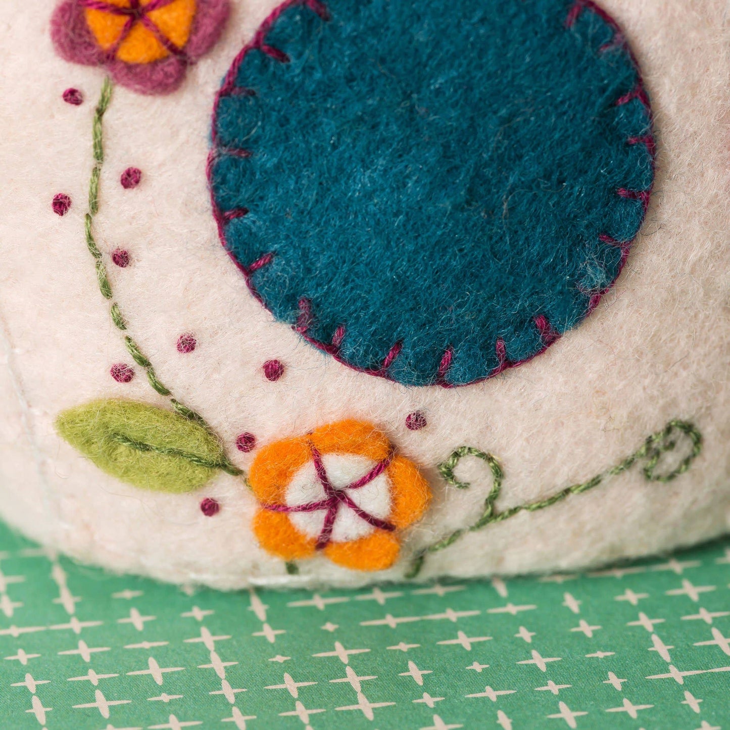 Close-up of a felted fabric piece from a bird house with embroidered flowers and a blue circle on a green and white checkered background.