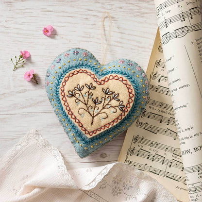 Embroidered heart-shaped pin cushion on a wooden surface with flowers and sheet music.
