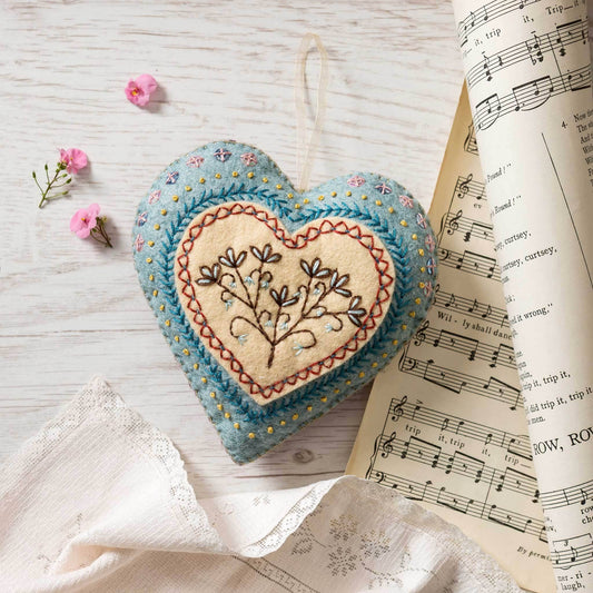 Embroidered heart-shaped pin cushion on a wooden surface with flowers and sheet music.