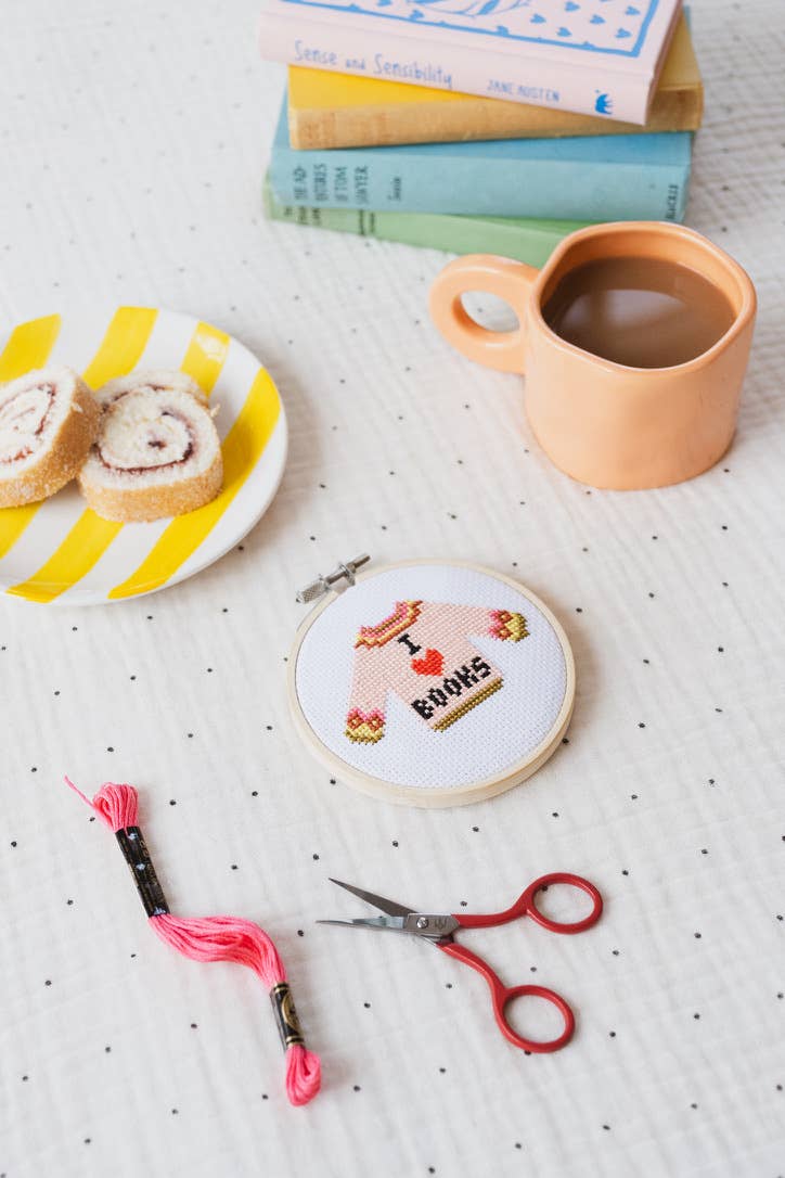 Cross-stitched hoop with 'I love books' design on a table with a mug, plate, and books.