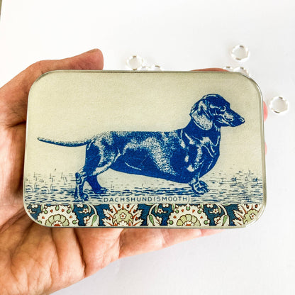 Hand holding a tin with a blue dachshund illustration on a white background