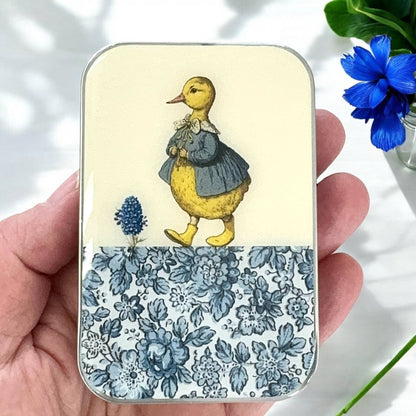 Small tin box with a duck illustration and floral design held by a hand.