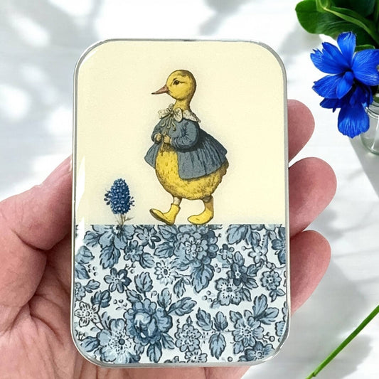 Small tin box with a duck illustration and floral design held by a hand.