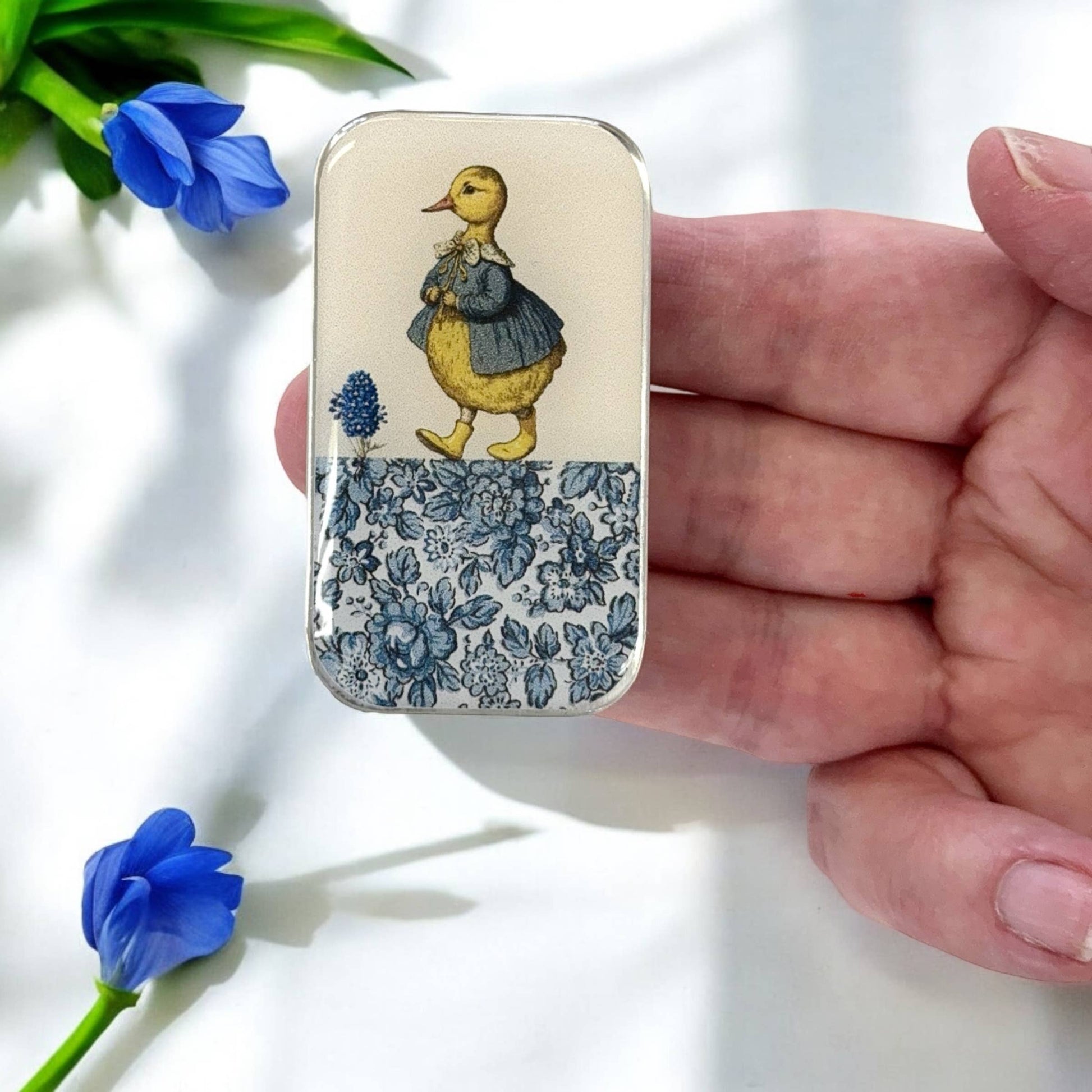 Small tin with a duck illustration held by a hand, with blue flowers in the background