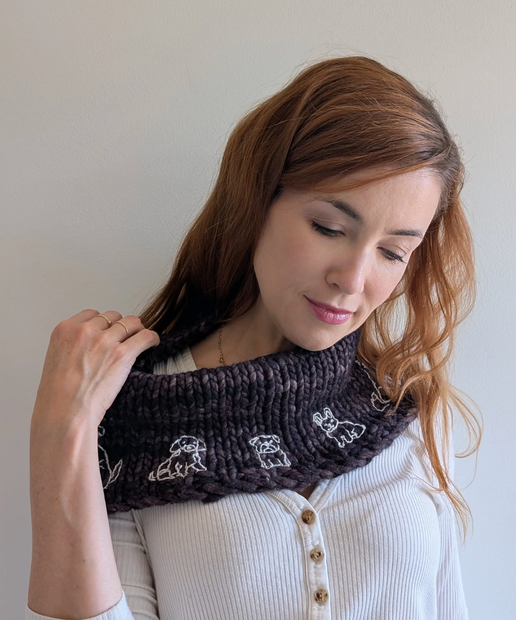 Woman wearing a dark knitted cowl with dog patterns against a plain background