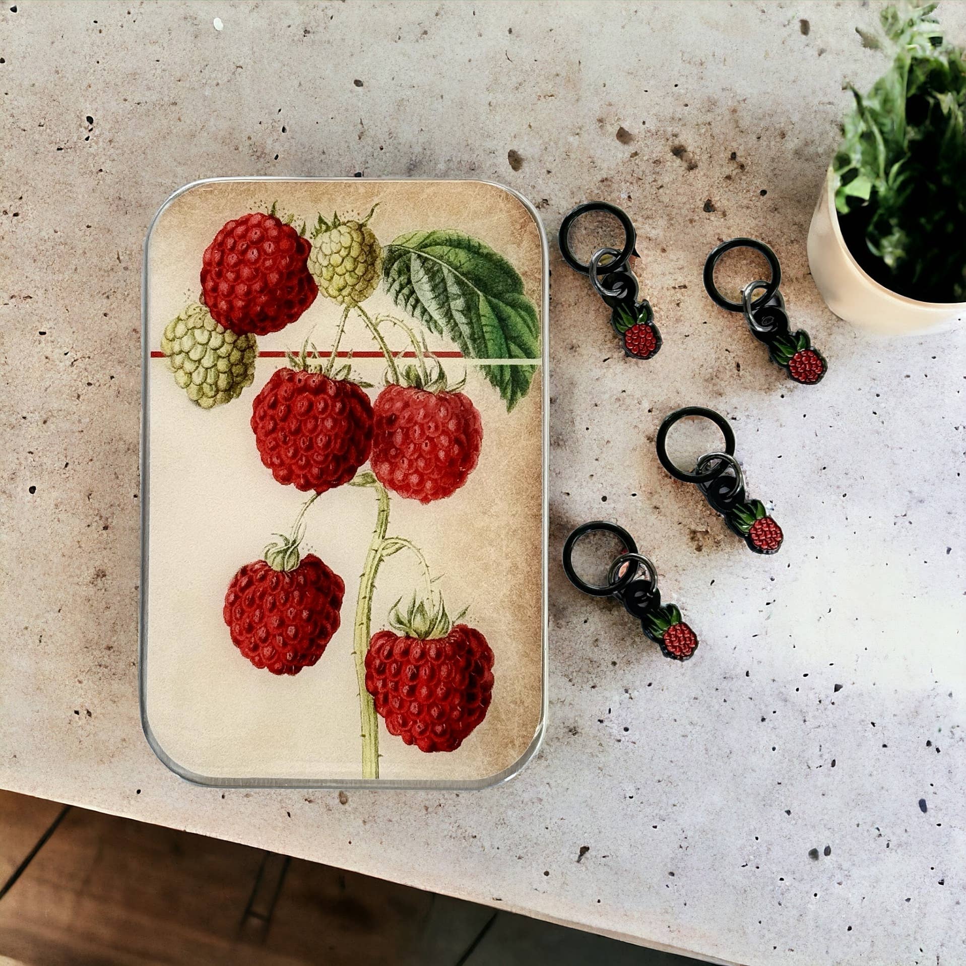 Decorative tin with raspberry design and matching keychains on a textured surface.